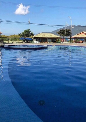 A swimming pool with a tiled deck, surrounded by buildings and a mountain in the background. - Arraial Praia dos Anjos Residence Club (Arraial do Cabo)