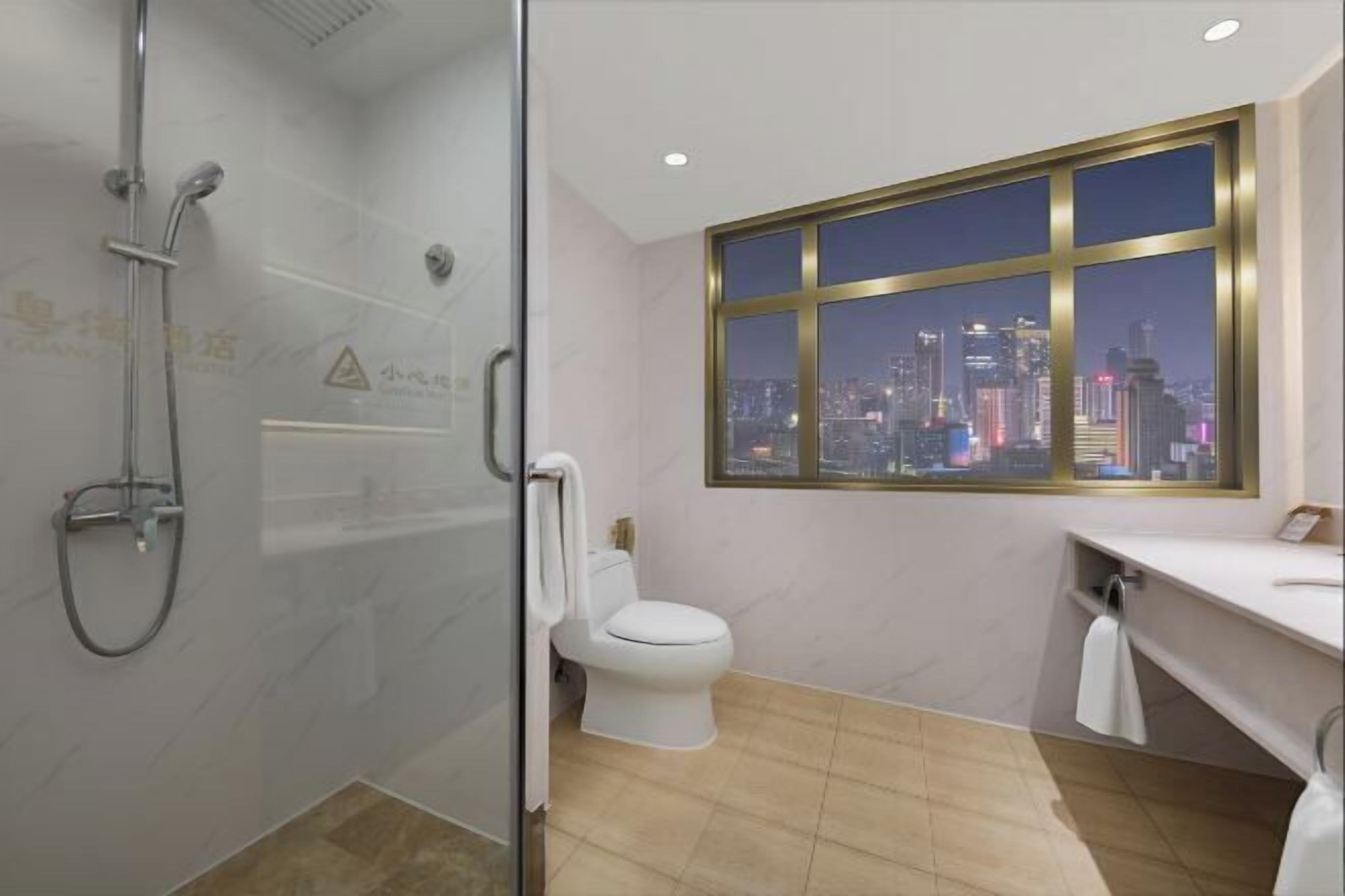 A bathroom with a glass shower enclosure, a toilet, a sink with a towel, and a window with a cityscape view.