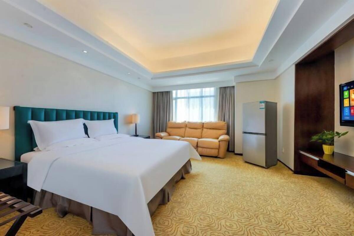A hotel room with a bed, a sofa, a TV, a refrigerator, and a window with curtains.