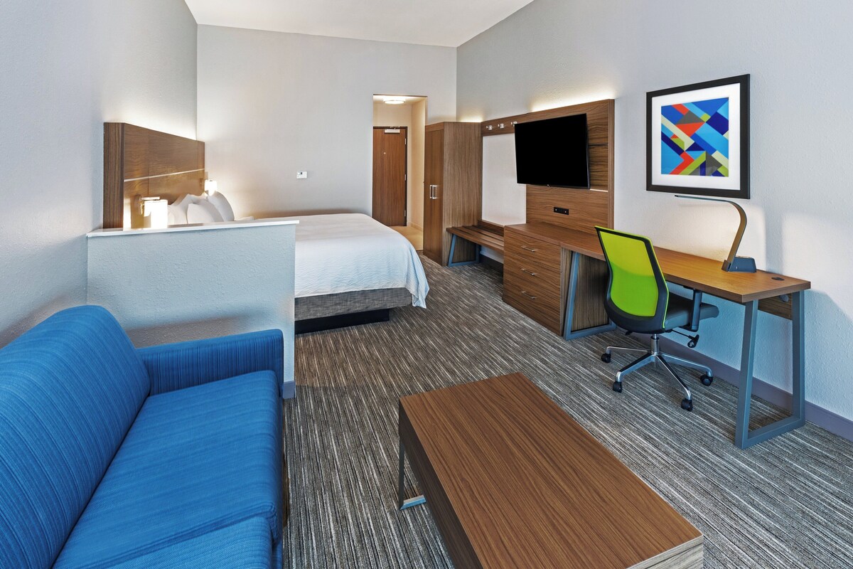 A hotel room with a bed, a blue sofa, a wooden desk with a chair, a flat-screen TV, and a framed abstract art piece on the wall.