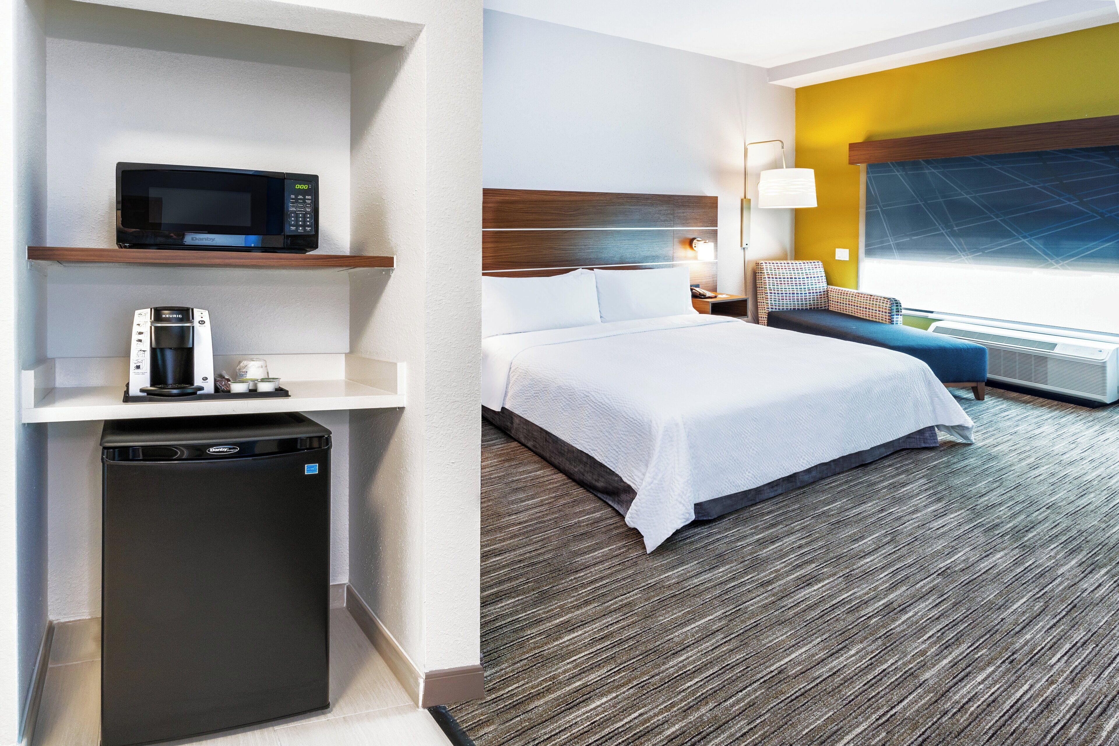 A hotel room with a bed, a microwave, a coffee maker, a small refrigerator, a bedside table with a lamp, and a window with a view.