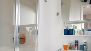 Une salle de bain avec une porte de douche en verre, un meuble-lavabo blanc avec un miroir et des articles de toilette posés sur le plan de travail.