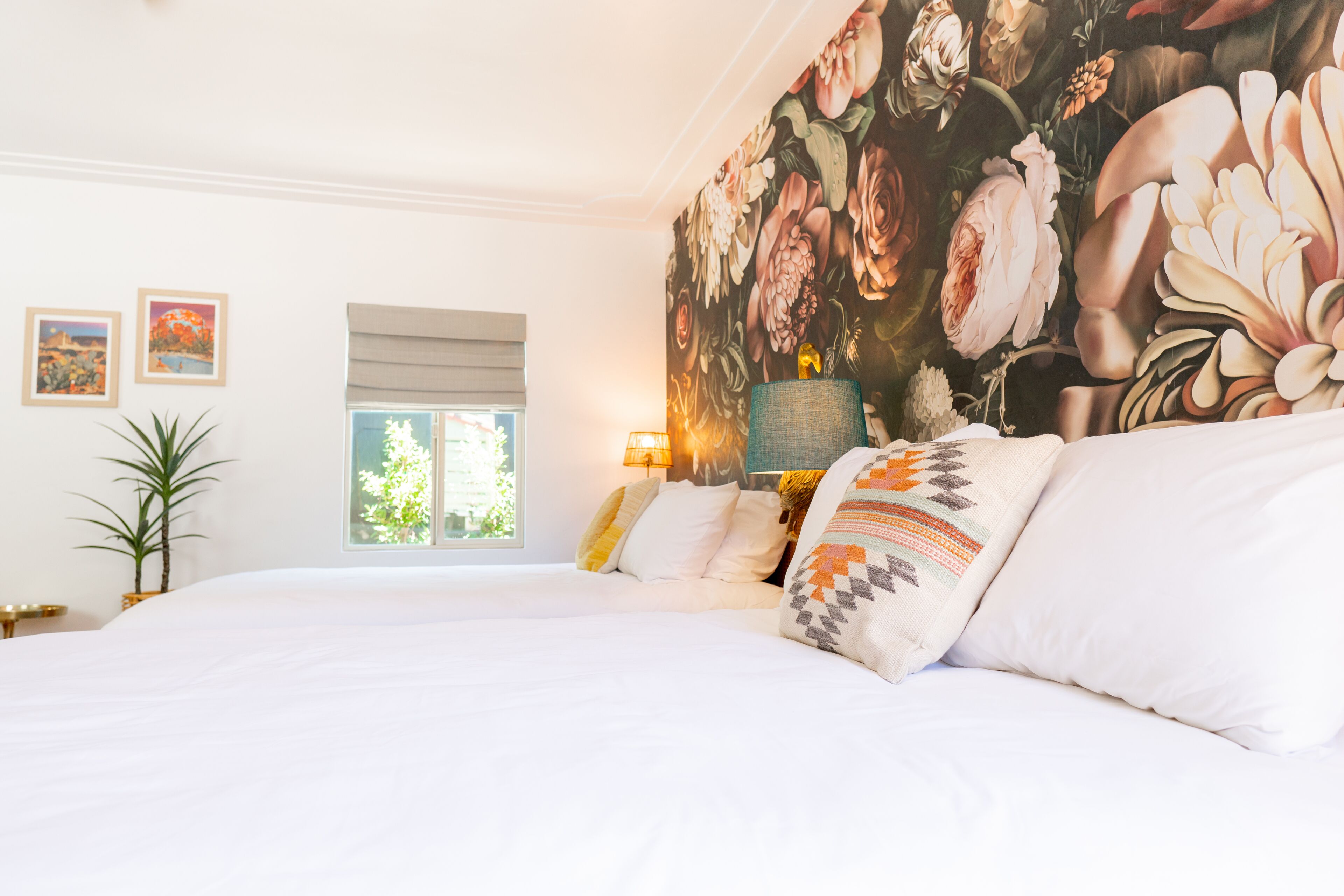 A bedroom with a large bed, a floral wall mural, a window with blinds, and a bedside lamp.