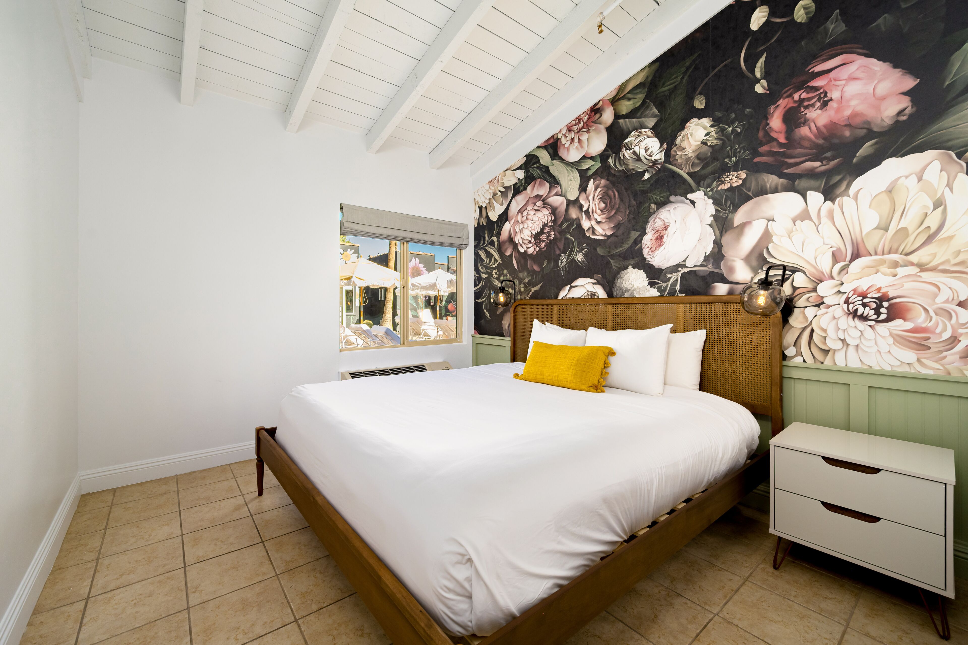 Une chambre avec un grand lit, une table de chevet, une fenêtre et une fresque florale sur le mur.