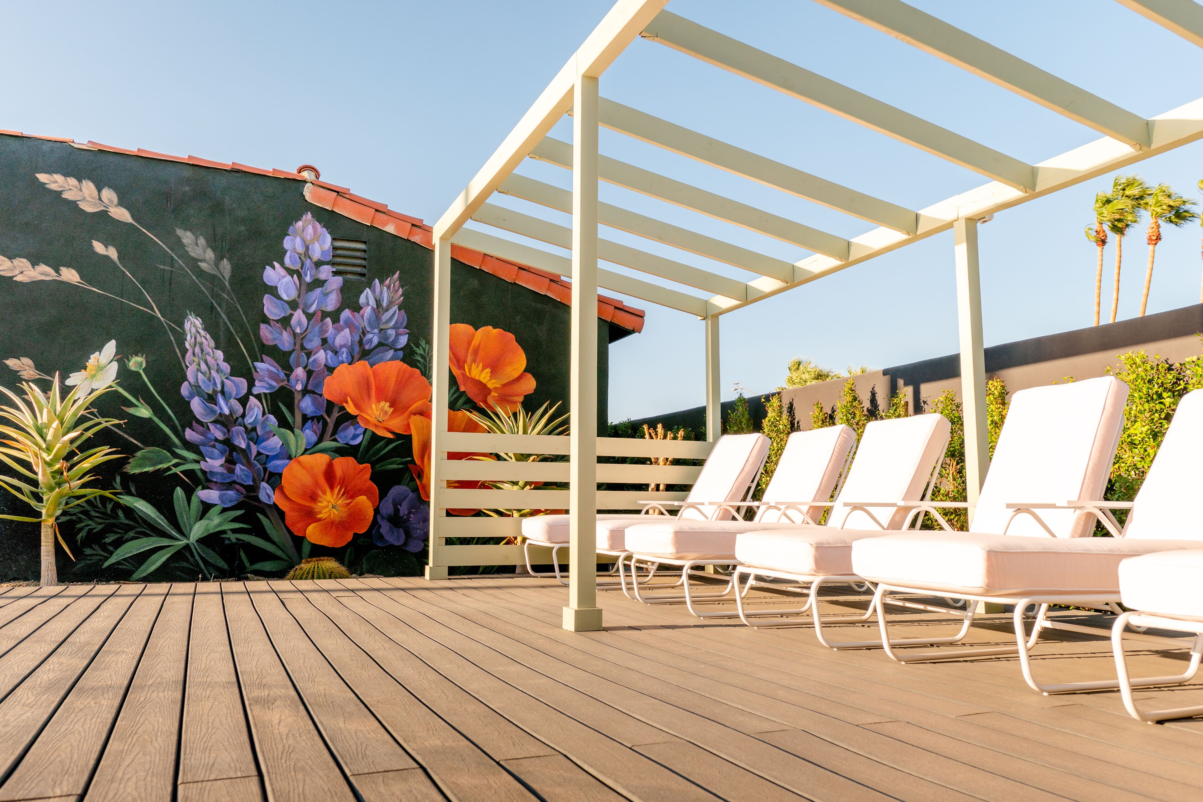 Eine Holzterrasse mit weißen Liegestühlen unter einer Pergola, eine Blumenmalerei und ein klarer Himmel.