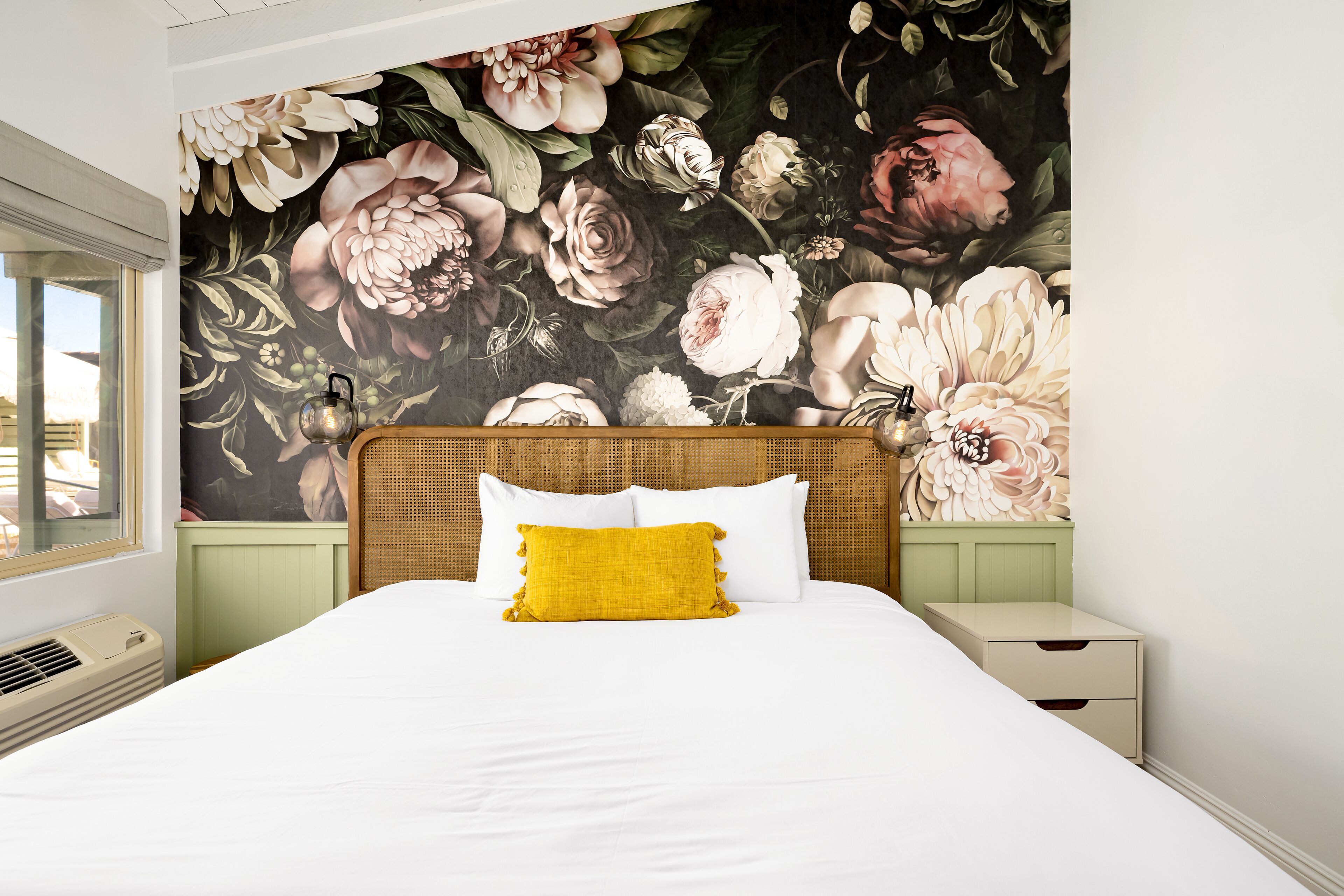 A bedroom with a large floral mural on the wall, a bed with white bedding and a yellow pillow, and a nightstand with drawers.