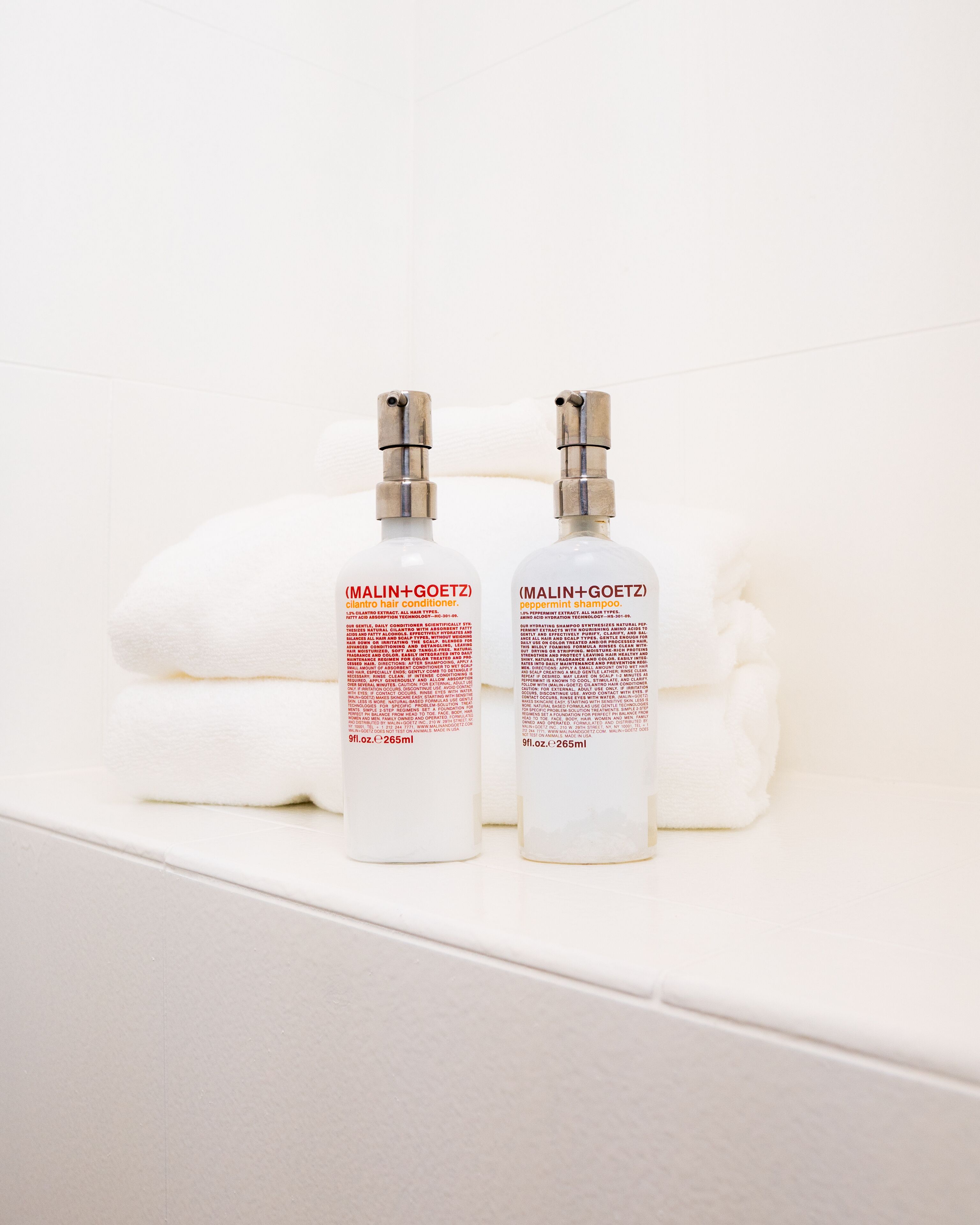Twee flessen MALIN+GOETZ haarconditioner en pepermuntshampoo op een wit oppervlak met opgevouwen handdoeken.