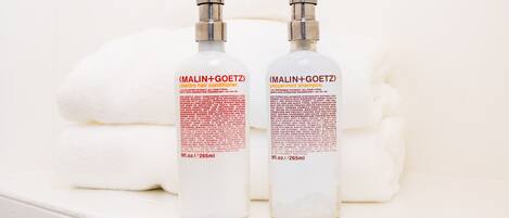 Twee flessen MALIN+GOETZ haarconditioner en pepermuntshampoo op een wit oppervlak met opgevouwen handdoeken.