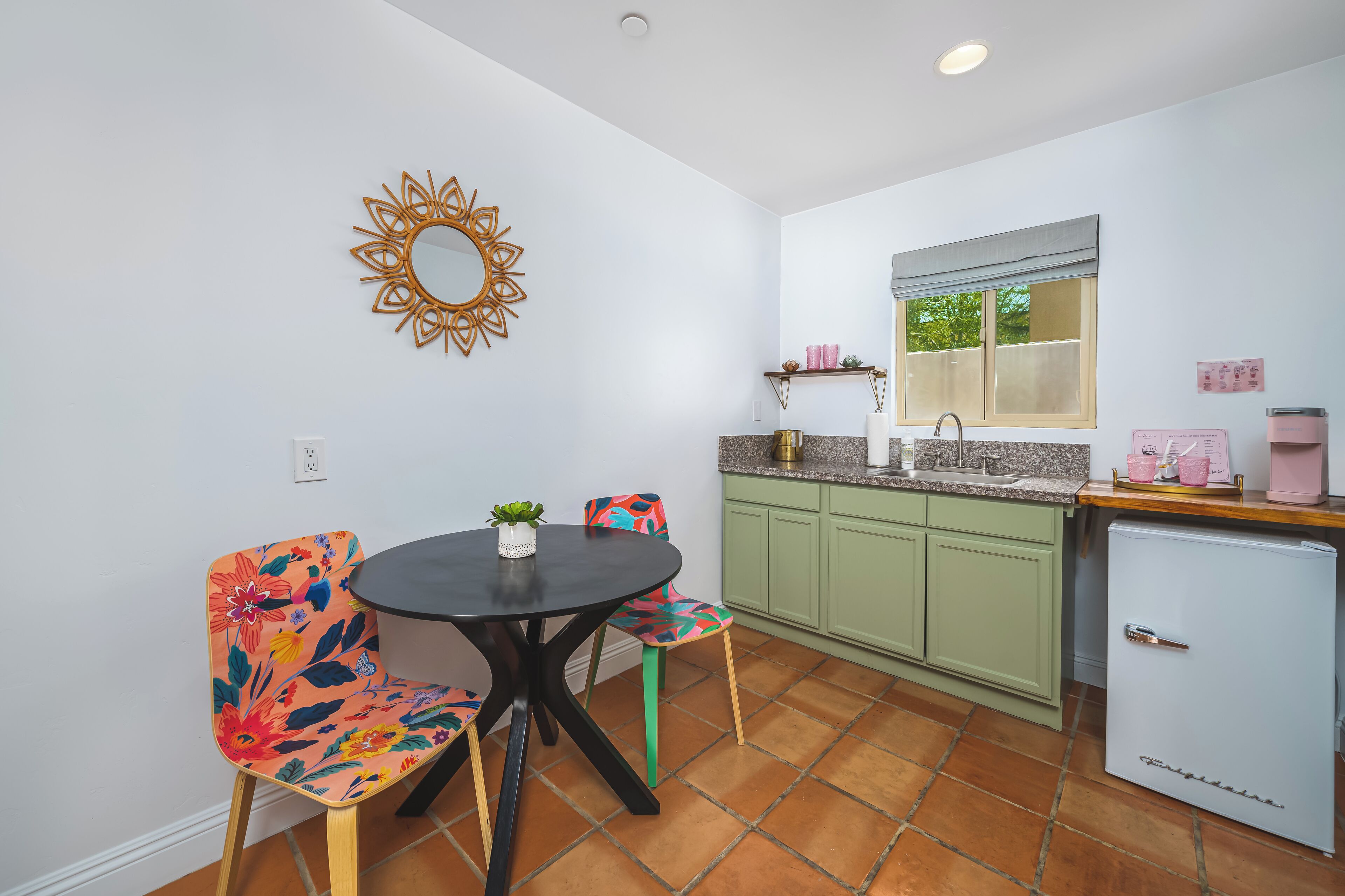 A compact kitchen with a dining area, featuring a round table, colorful chairs, a small refrigerator, and a window with a view.