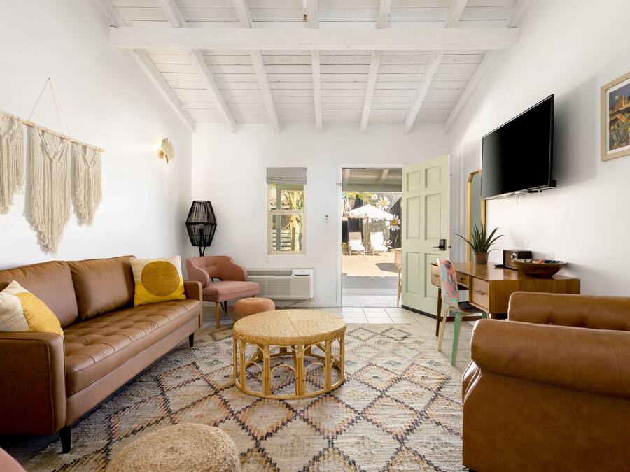A living room with a brown sofa, a round wicker ottoman, a small table, and a television on the wall.