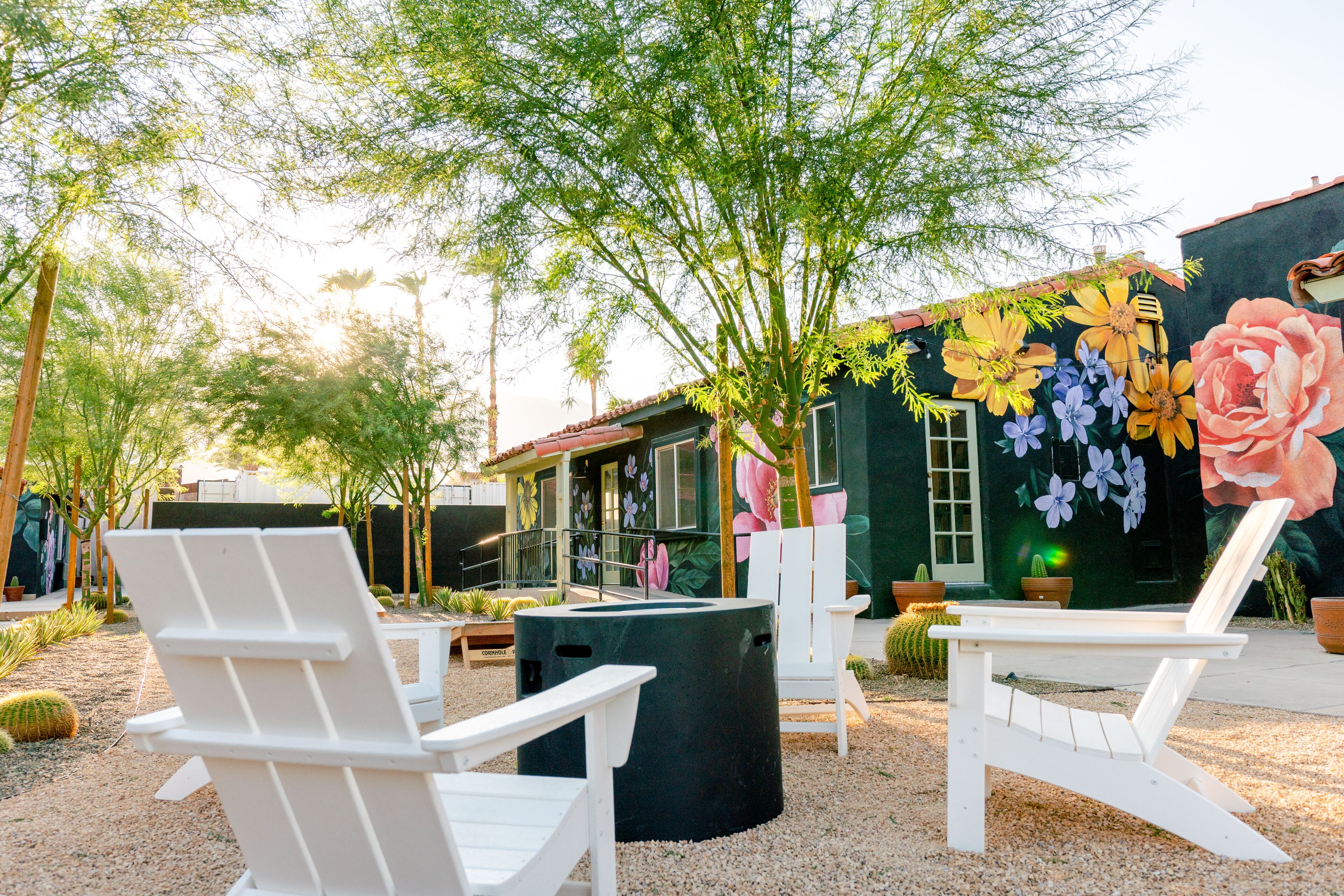 An outdoor seating area with white chairs and a black trash can, surrounded by cacti and a mural of flowers on a building.