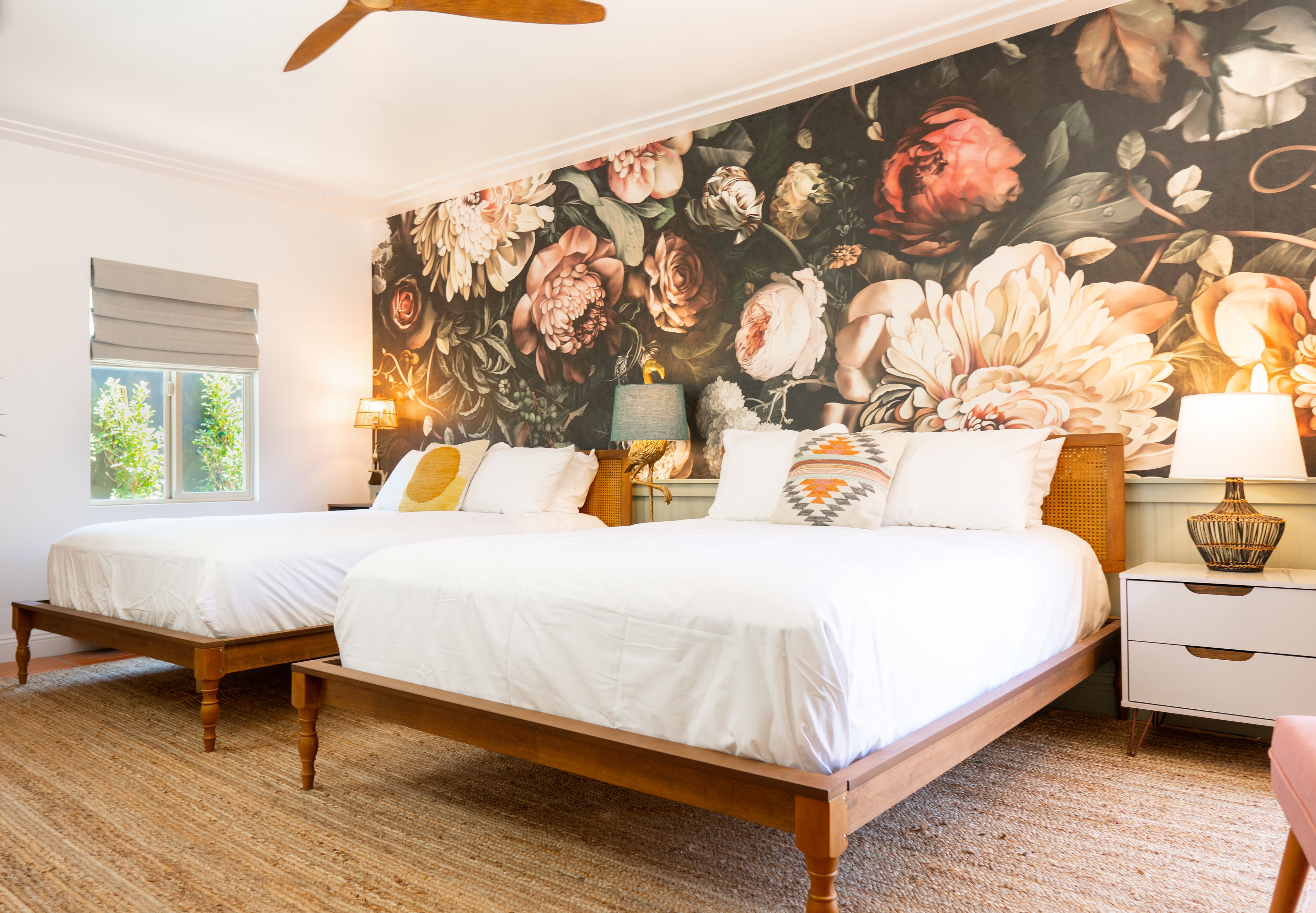 A bedroom with two wooden beds, a large floral mural on the wall, a window with blinds, and a nightstand with a lamp.