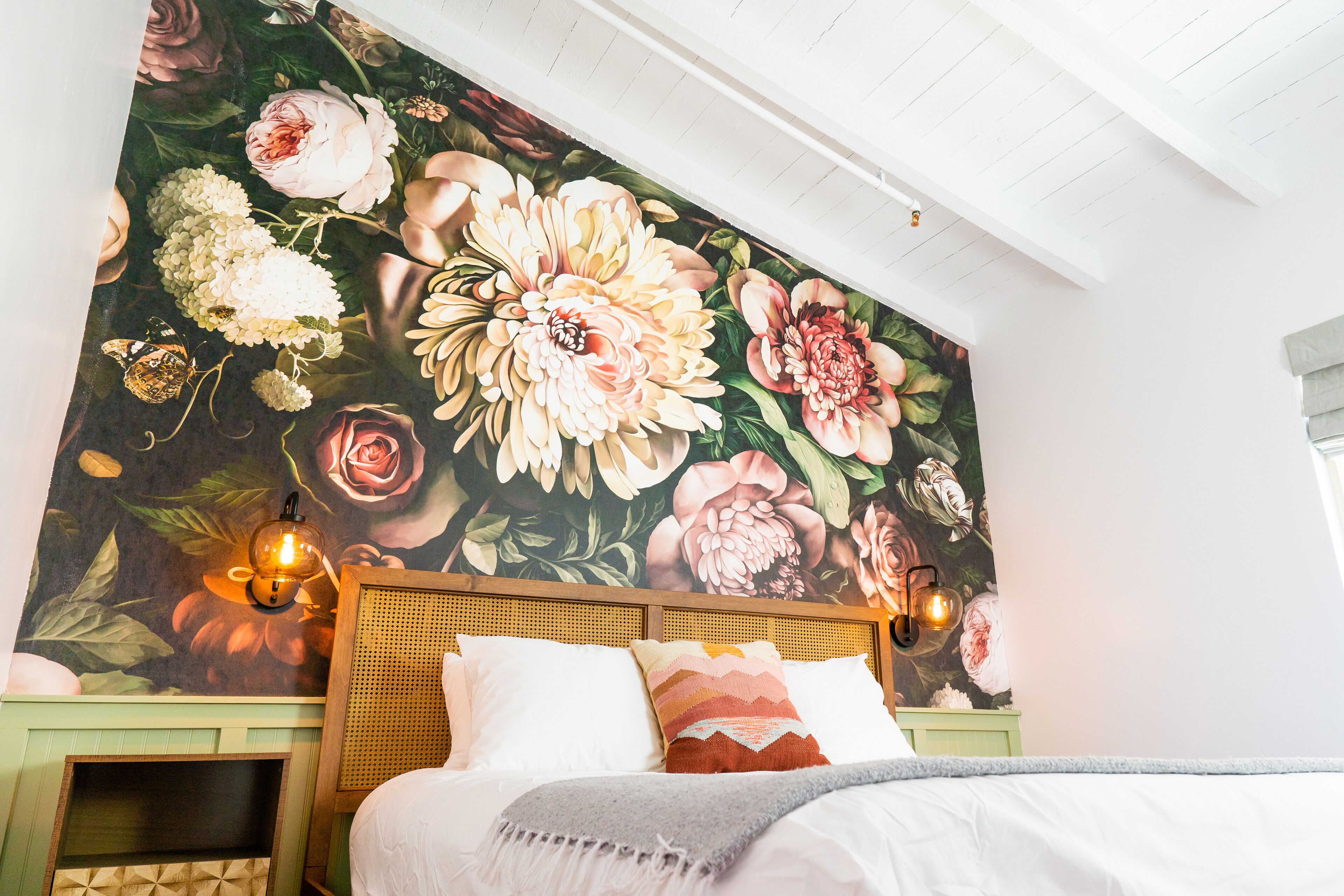 A bedroom with a large floral mural, a wooden headboard, two wall-mounted lamps, and a bed with white bedding and a gray throw blanket.