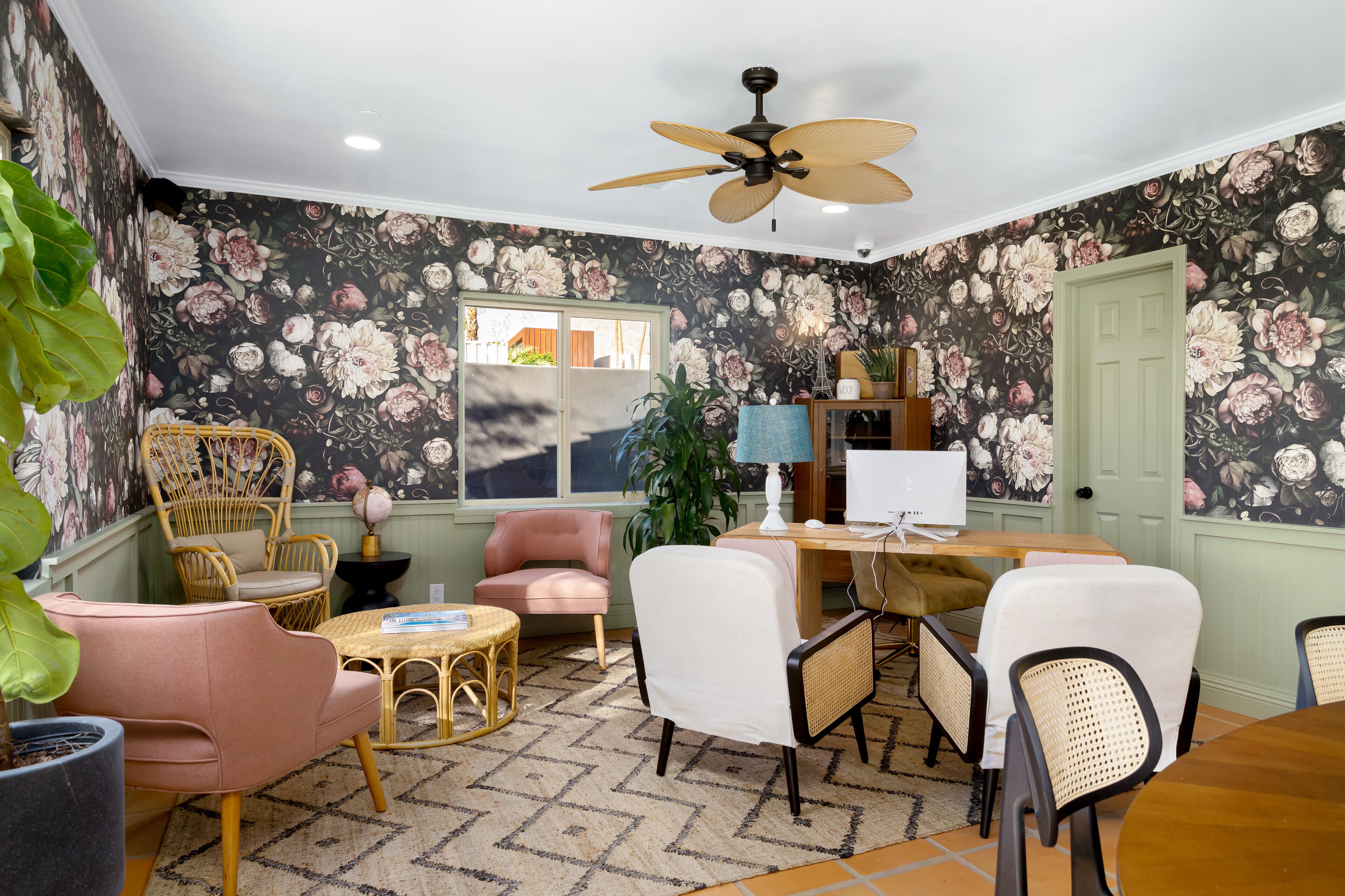 Een kamer met bloemen behang, een plafondventilator, een eettafel met stoelen en een raam met uitzicht.