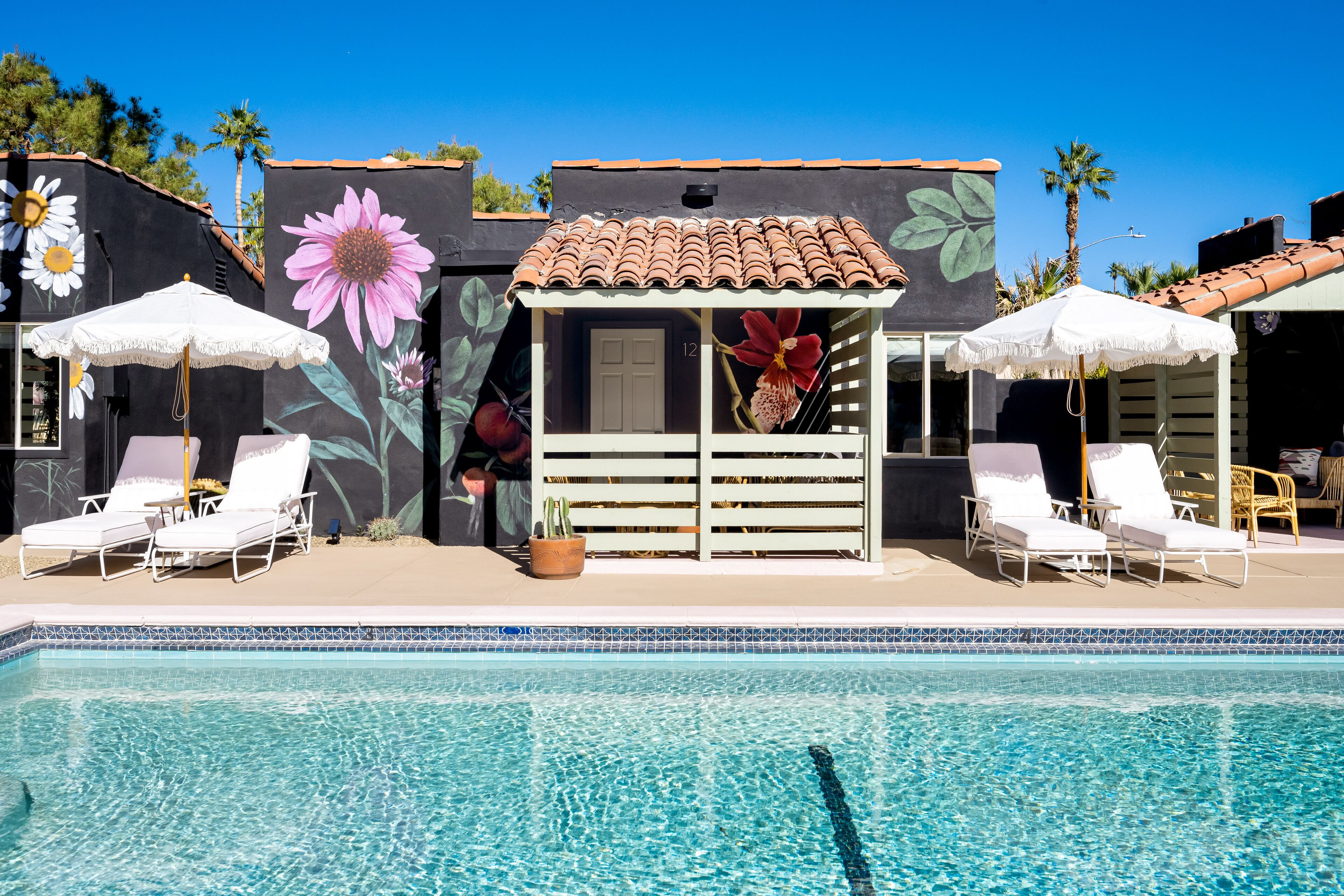 Área ao redor da piscina com espreguiçadeiras e guarda-sóis, telhado telhado e um edifício com murais florais.