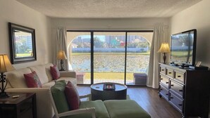 A hotel room with a sofa, a green ottoman, a TV, a lamp, and a view of a body of water.