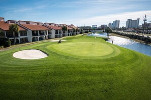 Um campo de golfe com lagoa, bunker de areia e um edifício com telhado vermelho ao fundo.