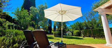 Une terrasse avec deux fauteuils en osier, un parasol blanc et un chemin en pierre.