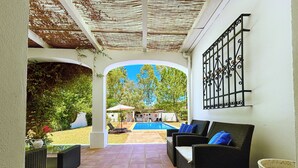 Un patio techado con mobiliario de mimbre, una piscina y vistas al jardín.