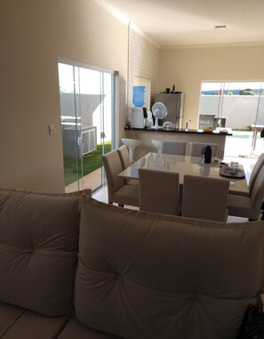 A modern living room with a glass dining table, beige sofas, and a kitchen area with a refrigerator and a water dispenser.