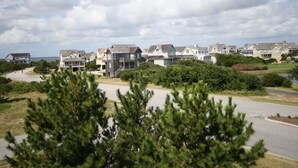 A coastal residential area with multiple two-story houses, a paved road, and a body of water in the background. - Lauren Lee: Private Pool, Hot Tub, Golf Discounts (Nags Head)
