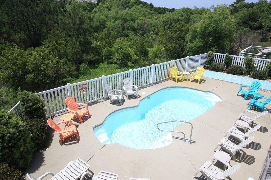 A swimming pool surrounded by white and yellow Adirondack chairs, with a white fence and lush greenery in the background.