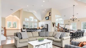 A spacious living room with a sectional sofa, a coffee table, and a dining area with a chandelier. - 8th And Sound: Elevator | Pool | Hot Tub (Corolla)
