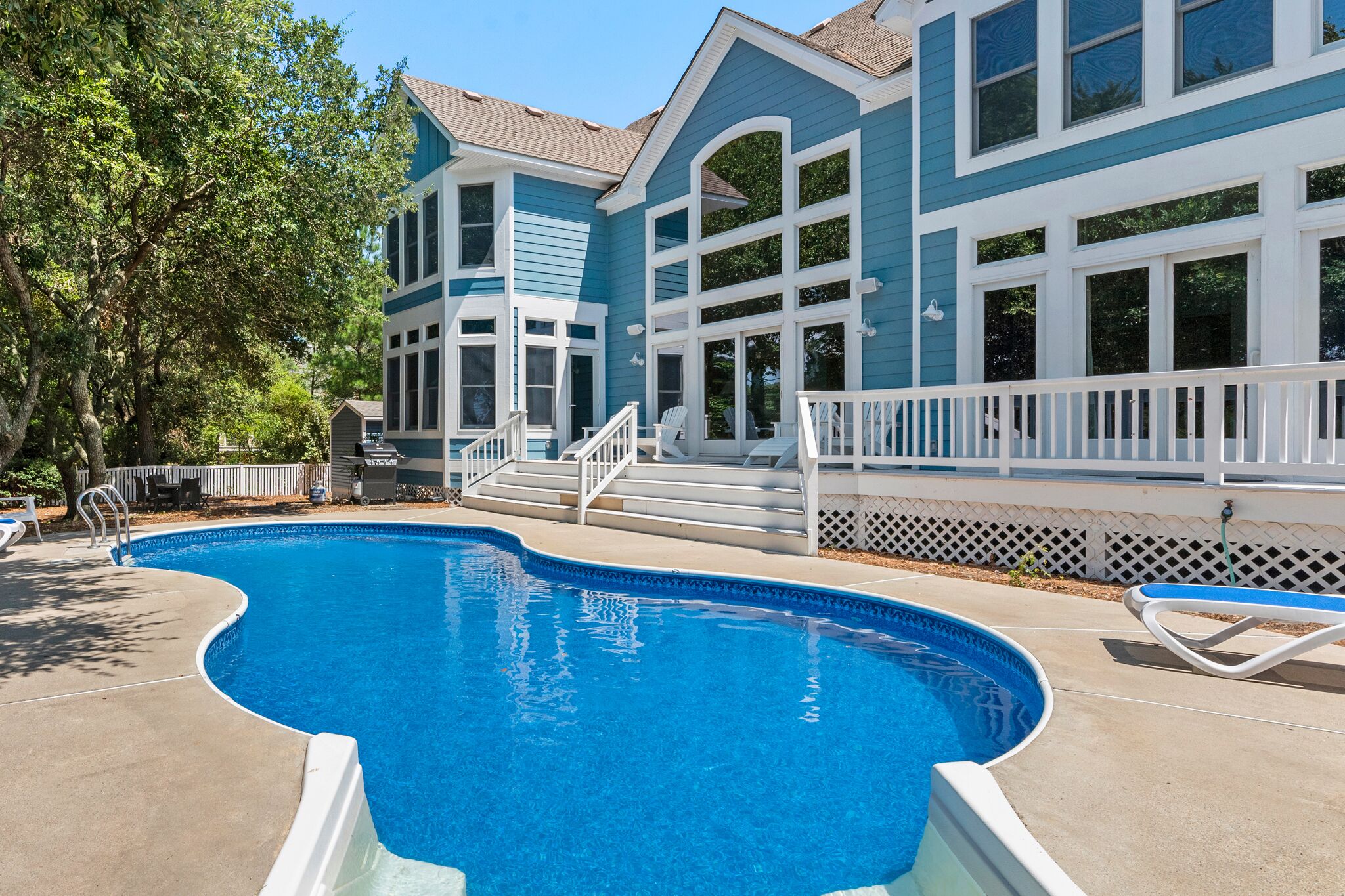 Ein Pool mit gebogener Form, umgeben von einer Terrasse mit weißen Geländern und Stufen, die zu einem Gebäude mit großen Fenstern und blauer Fassade führen.