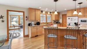 A modern kitchen with wooden cabinets, a central island with bar stools, pendant lighting, and a view to the dining area. - 40-Love is a:  Soundfront | Private Pool | Spa (Corolla)