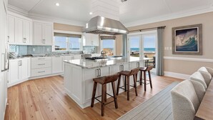 Une cuisine spacieuse avec des meubles blancs, un grand îlot central doté de tabourets et une vue sur l’océan grâce à de grandes fenêtres.