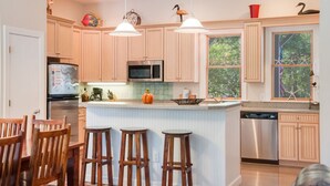 A kitchen with wooden cabinets, a central island with stools, stainless steel appliances, and a window with a view of trees. - Reminiscent: Oceanside, Private Pool, Spa (Nags Head)