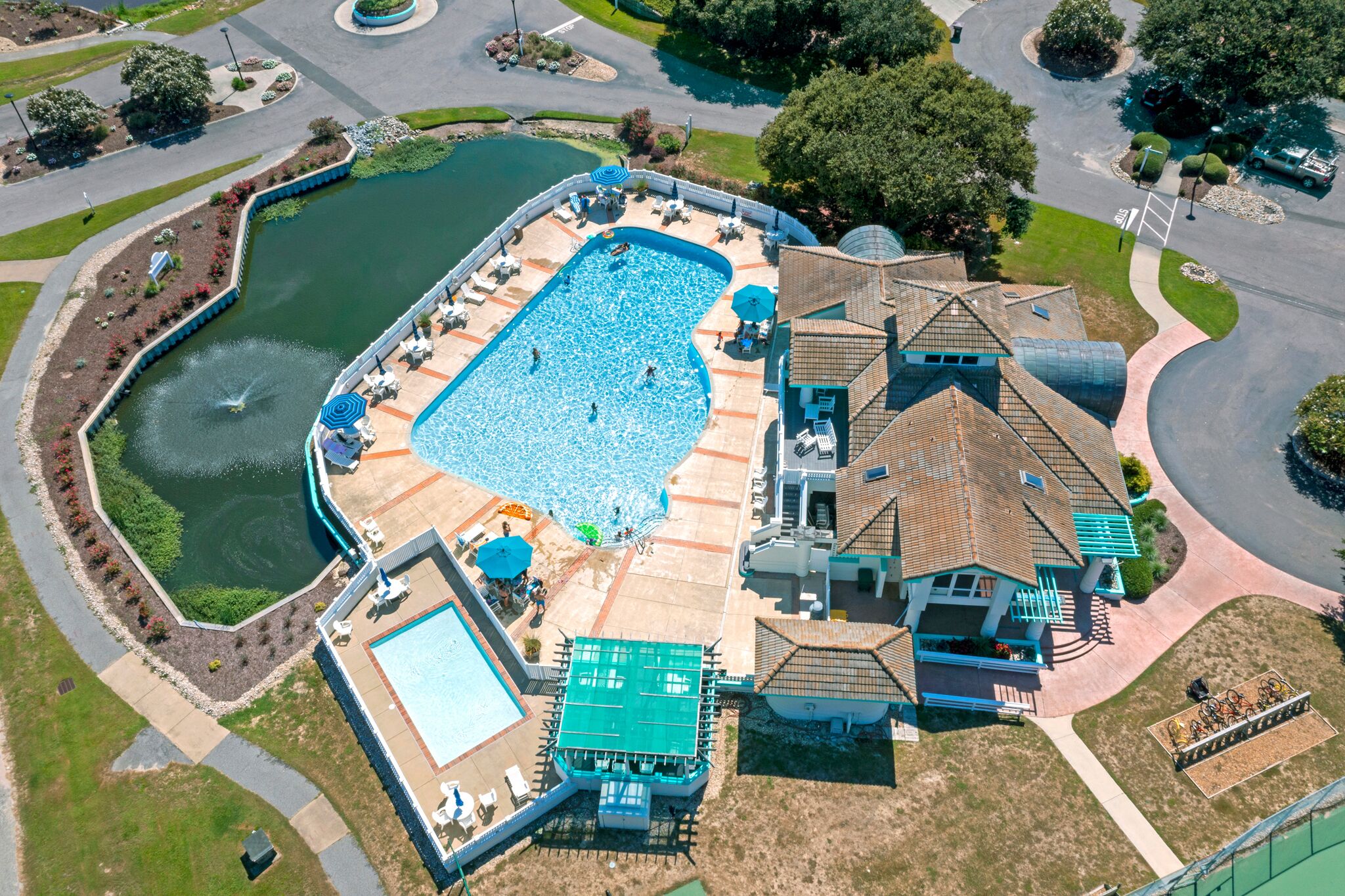 Vue aérienne d’un quartier résidentiel avec une grande piscine, un court de tennis et une maison dont le toit présente un design original.