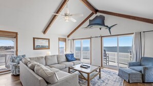 A spacious living room with a large sofa, a coffee table, and a view of the ocean through large windows. - All Decked Out: Soundfront | Rooftop Decks (Nags Head)
