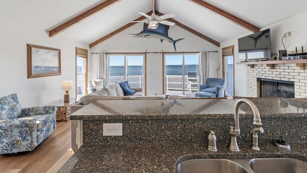 A kitchen with a large island, wooden beams, a ceiling fan, and a view of the ocean. - All Decked Out: Soundfront | Rooftop Decks (Nags Head)