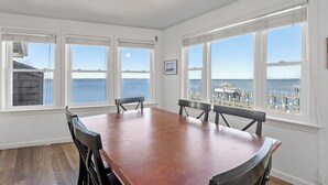 A dining room with a large table and chairs, large windows with ocean views, and a ceiling fan. - All Decked Out: Soundfront | Rooftop Decks (Nags Head)