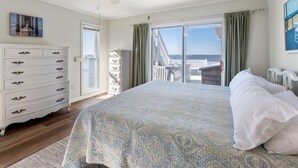 A bedroom with a large bed, a ceiling fan, a dresser, and a view of the sea through a sliding door. - All Decked Out: Soundfront | Rooftop Decks (Nags Head)