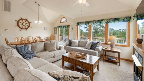 Un salon spacieux avec un grand canapé d’angle, une table basse en bois et un ventilateur de plafond. On y trouve également un coin repas avec des chaises et une table en bois, ainsi que de grandes fenêtres avec des rideaux.