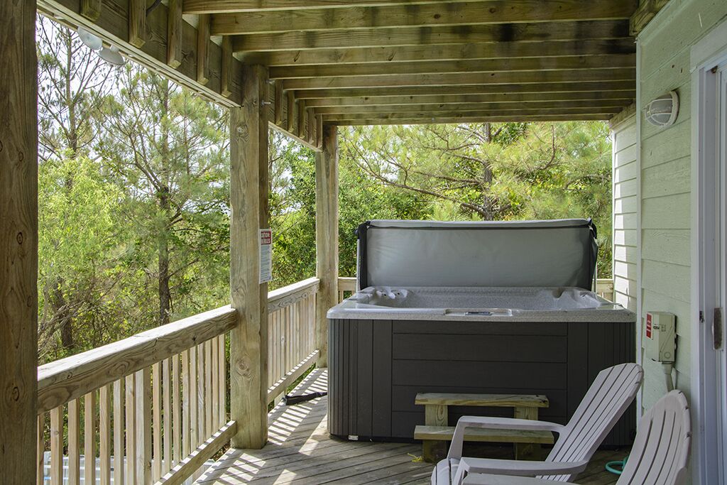 A covered outdoor deck with a hot tub, wooden chairs, and a view of trees.