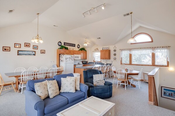 Uma sala de estar espaçosa com um sofá azul, área de jantar com cadeiras brancas, uma cozinha com armários de madeira e uma placa com a inscrição "BEACH HOUSE".