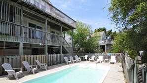 A pool area with a wooden deck, white lounge chairs, and a two-story building with balconies. - A Dolphin's Cove: Rec Room w/ Pool Table, Private Pool & Spa, Dogs OK! (Corolla)