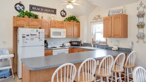 A kitchen with wooden cabinets, a white refrigerator, a microwave, a stove, and a large island with bar stools. There is a ceiling fan, a sign that says "BEACH HOUSE," and decorative plates on the wall. - A Dolphin's Cove: Rec Room w/ Pool Table, Private Pool & Spa, Dogs OK! (Corolla)