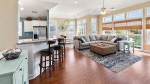 A spacious living area with a large sofa, a coffee table, and a dining area with chairs and a kitchen in the background. - Craba-Cabana: Surf, Sand, and a Private Pool; Community Pool and Tennis Court (Corolla)