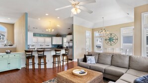 A spacious living room with a grey sofa, a woven coffee table, and a kitchen area with white cabinets and a bar stool. - Craba-Cabana: Surf, Sand, and a Private Pool; Community Pool and Tennis Court (Corolla)