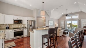 A spacious kitchen with white cabinetry, a central island, and a dining area with dark chairs and a wooden table. - A Ruff Life: Saltwater Pool, Hot Tub, 500 ft. to the Beach, Dog-friendly (Corolla)