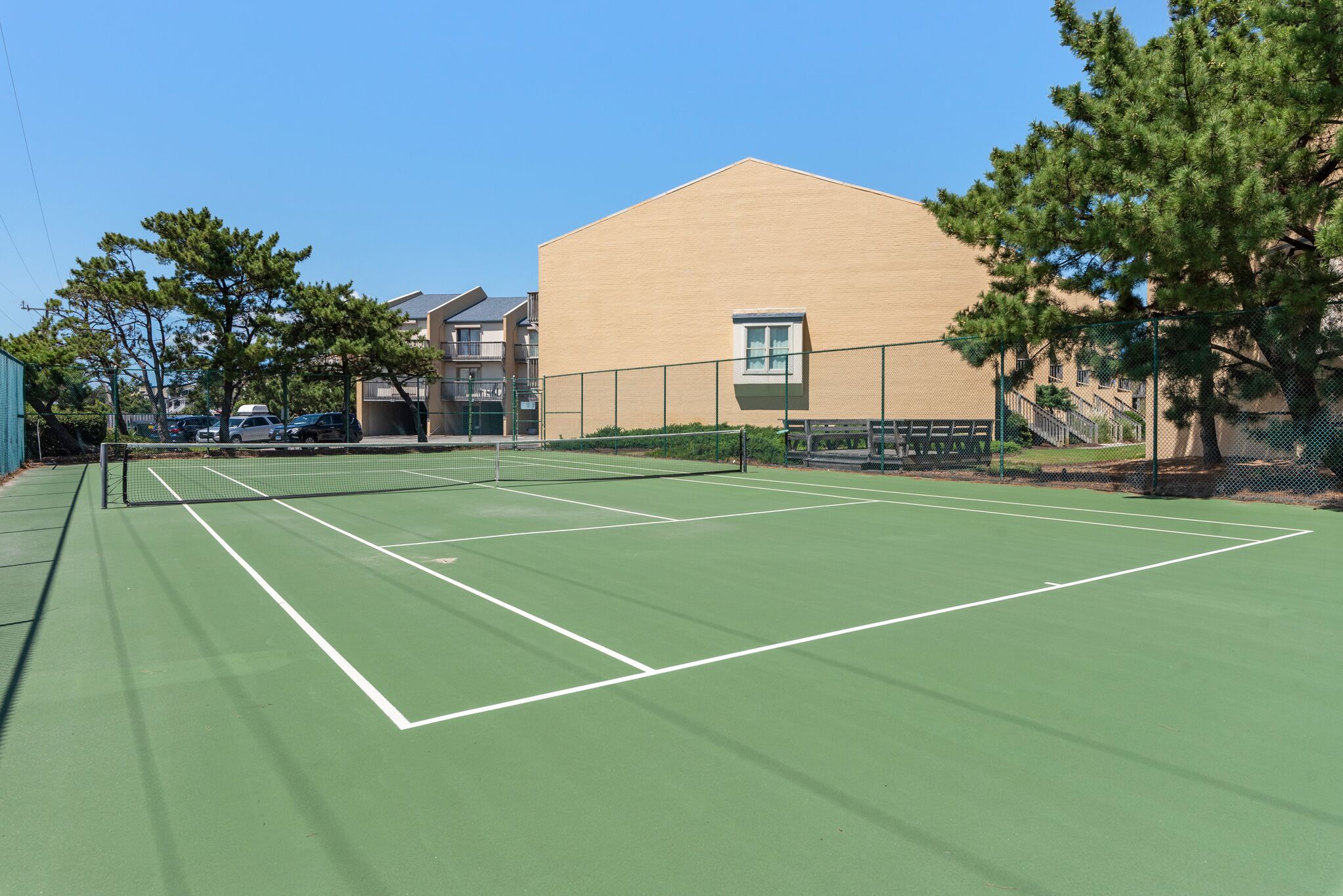 Ein Tennisplatz mit Netz, umzäunt und neben einem Gebäude mit Fenster und Tür.