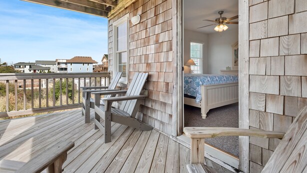 Um deck de madeira com área de estar externa, um quarto com cama e ventoinha no teto, e vista para casas vizinhas.