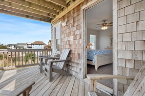 Une terrasse en bois avec coin salon extérieur, une chambre dotée d’un lit et d’un ventilateur de plafond, et une vue sur les maisons voisines.