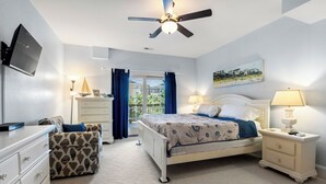 Ein Schlafzimmer mit Bett, Nachttisch, Kommode, Stuhl, Deckenventilator und einem Fenster mit Vorhängen.