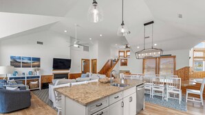 Amplia sala de estar con una isla grande, piso de madera y un comedor con sillas blancas y una mesa de madera.