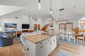 A spacious living area with a large island, wooden flooring, and a dining area with white chairs and a wooden table. - E=M(Sea)2: Private Pool & Hot Tub, Rooftop Deck w/ Ocean View, Pet-friendly (Corolla)